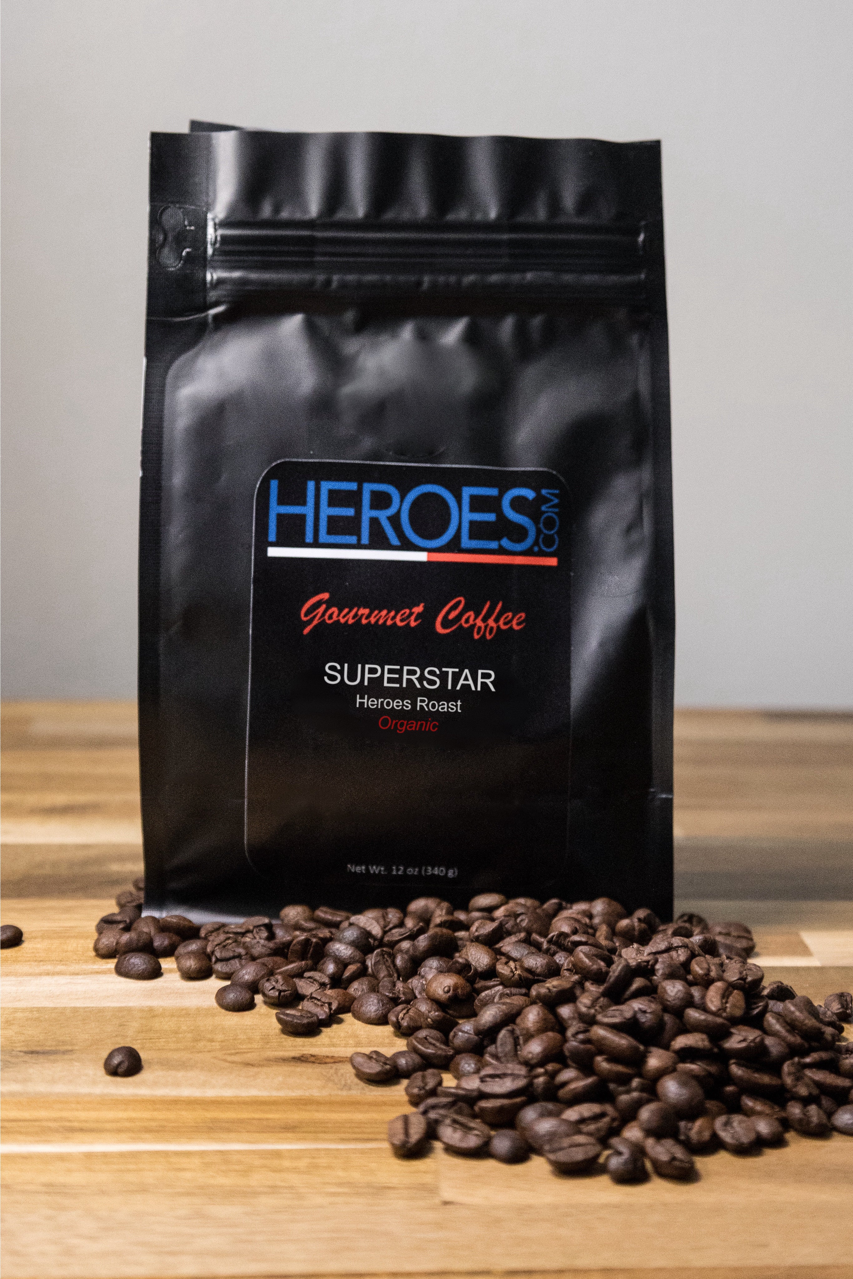 A black bag of Heroes Gourmet Coffee labeled Superstar Heroes Roast Organic sits on a wooden surface with coffee beans scattered in front of it.