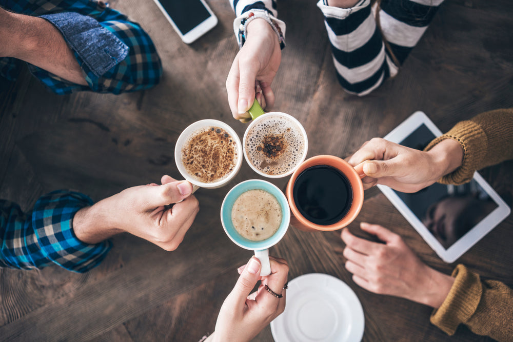 Four people holding different coffee mugs together in a toast over a wooden table, with a smartphone, a tablet, and a white saucer visible in the background.