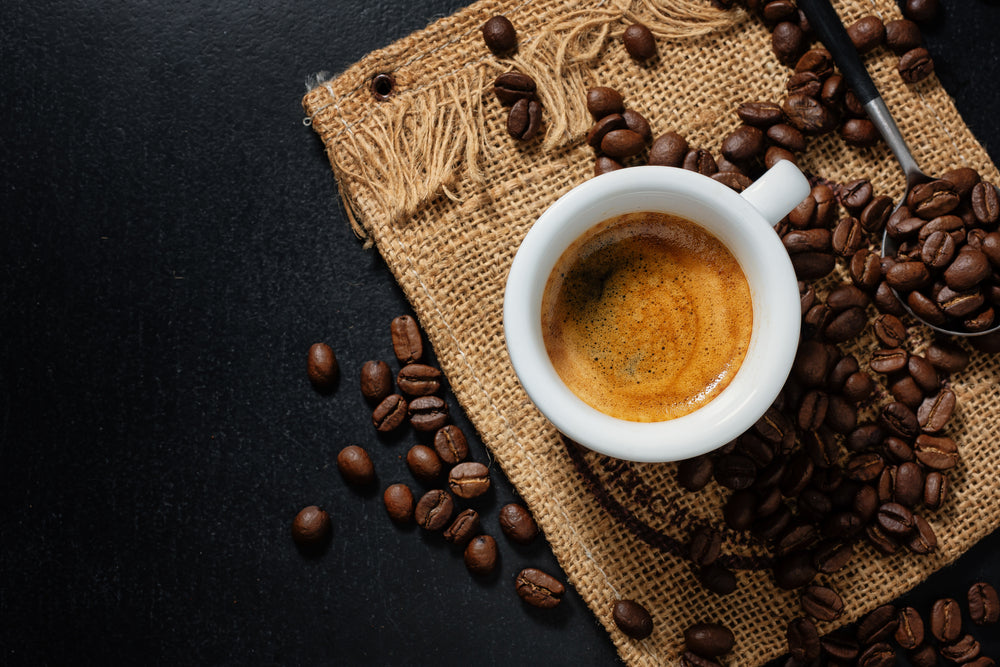 A white cup of espresso on a burlap mat, surrounded by scattered coffee beans, sits on a dark surface.