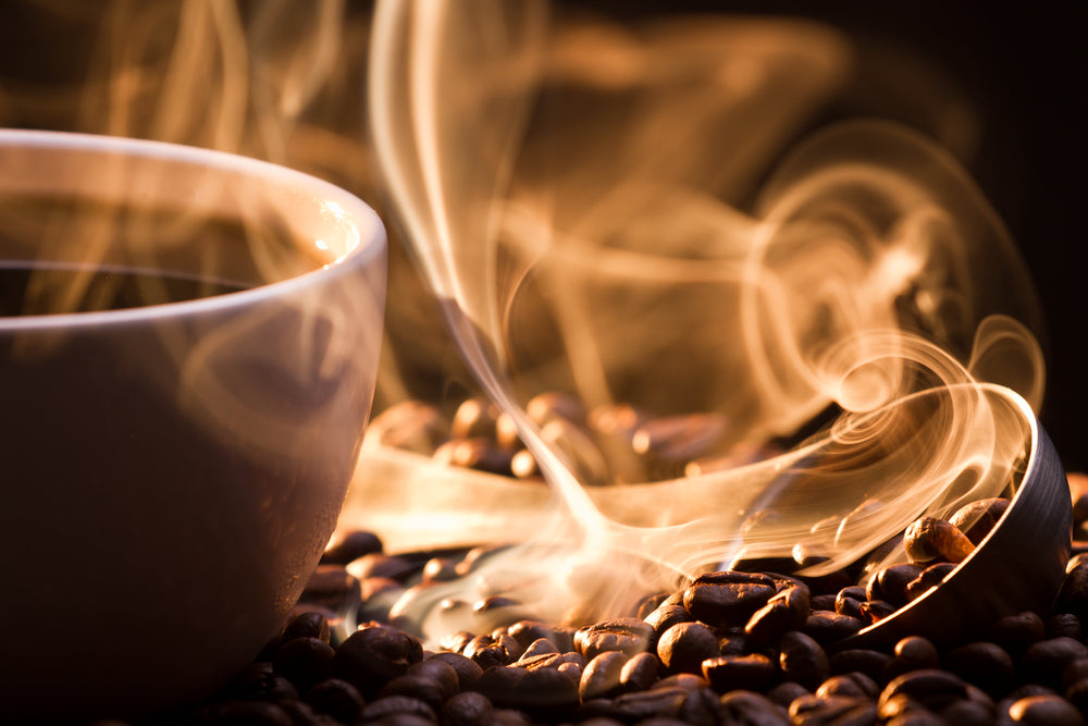 A steaming cup of coffee sits close to a pile of roasted coffee beans, with aromatic steam swirling and rising in the warm light.