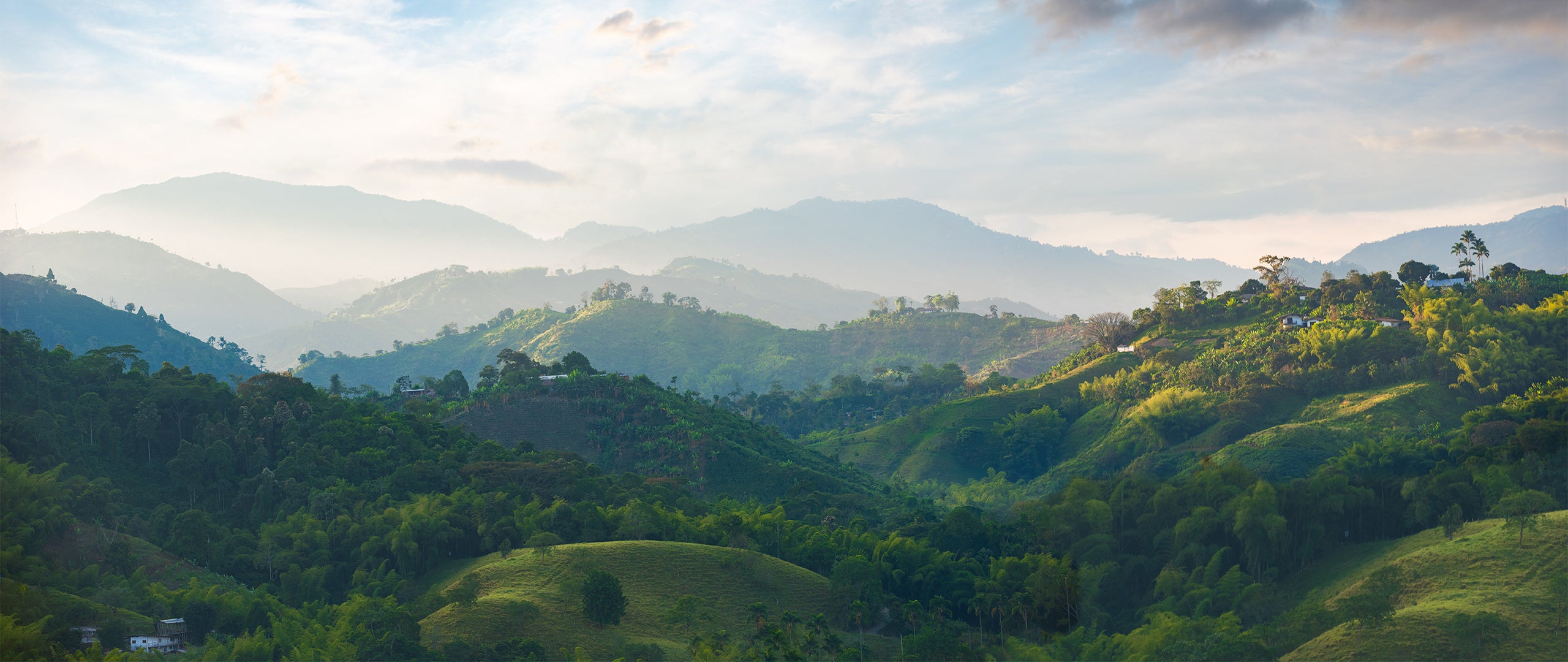 A panoramic view of lush green hills and valleys under a cloudy sky with soft sunlight, layers of distant mountains in the background, and scattered trees and small houses dotting the landscape.