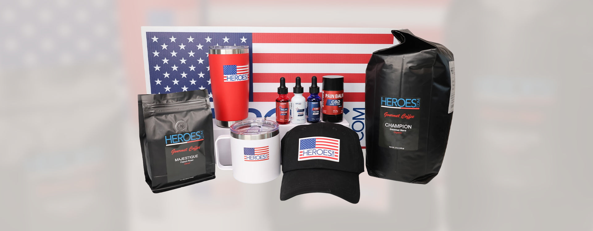 A display of HEROES coffee bags, red and white tumblers, health supplements, a black cap, and an American flag sign in the background. All items feature the HEROES brand logo and American flag design.