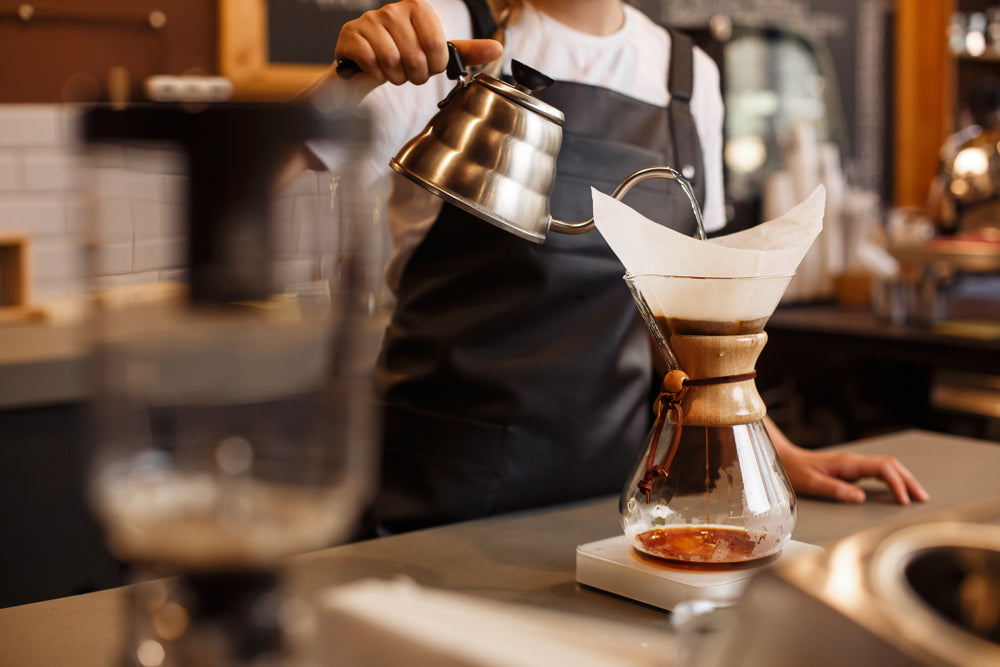 A barista wearing a black apron pours hot water from a gooseneck kettle into a Chemex coffeemaker on a scale, brewing coffee in a cozy café setting.