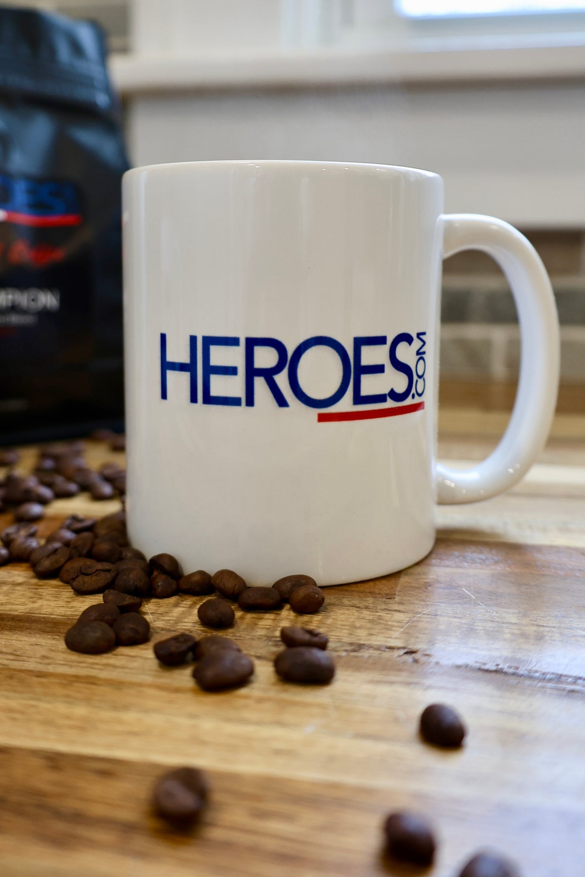 A white mug with the word HEROES.com in blue and red letters sits on a wooden surface, surrounded by scattered coffee beans. A blurry coffee bag is in the background.
