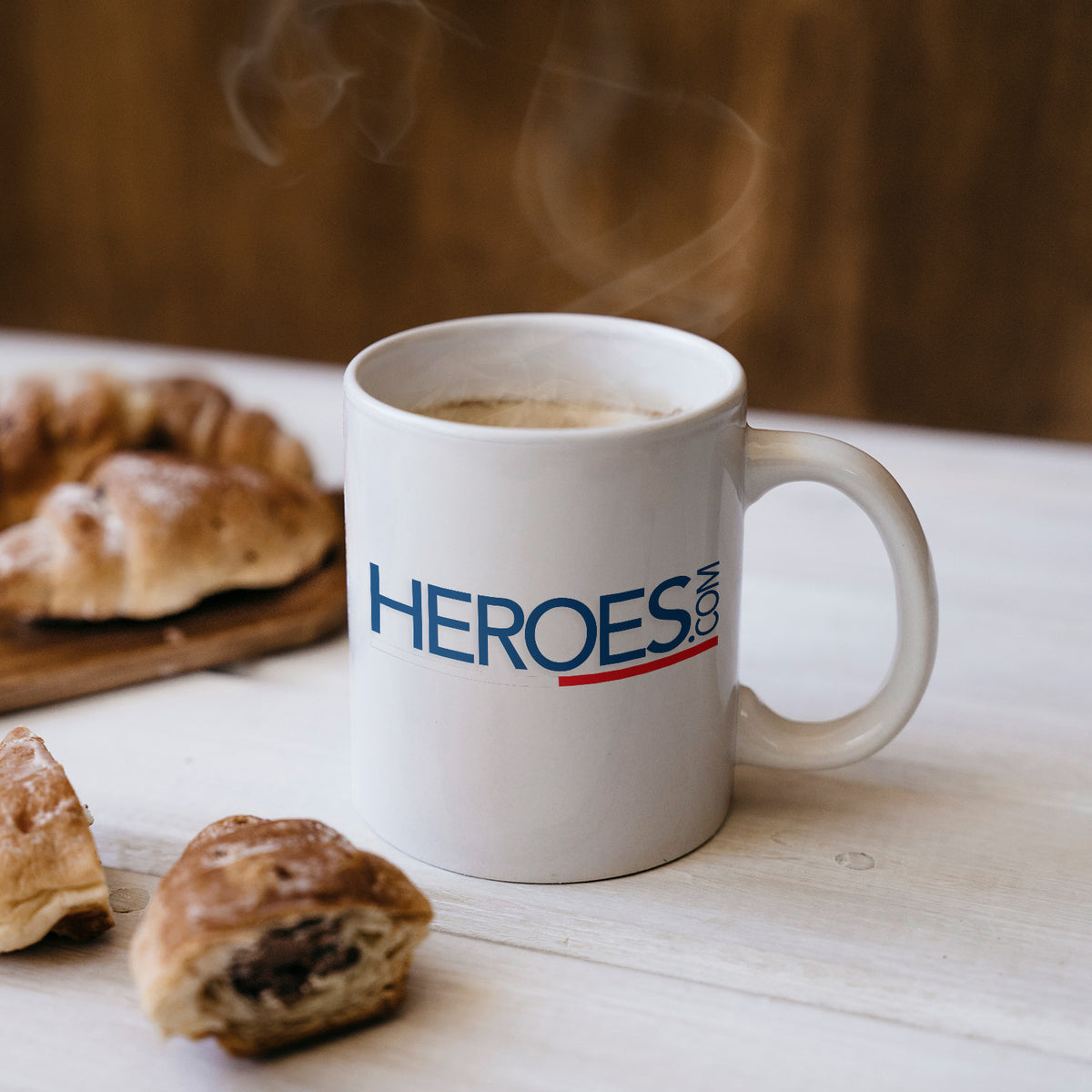 A white mug with the word HEROES.COM printed on it sits on a white table. There is steam rising from the mug, and pastries, including croissants and chocolate bread, are nearby.