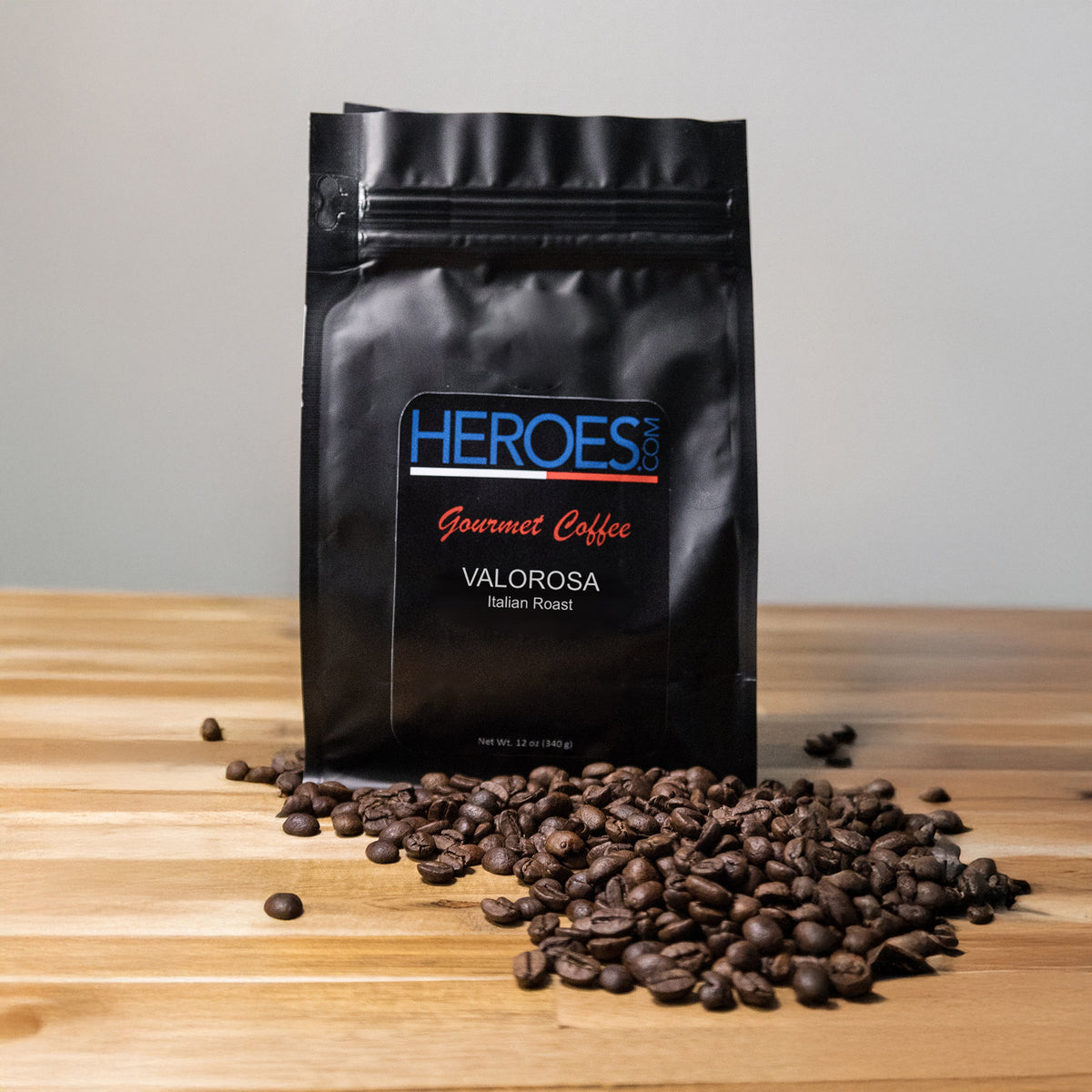 A bag of VALOROSA Italian Roast sits on a wooden surface with Arabica coffee beans scattered in front, set against a plain light background.